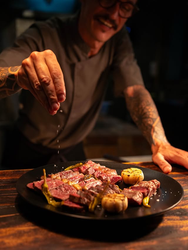 A chef sprinkling seasoning on a finished plate of gourmet steak from Chupacabras