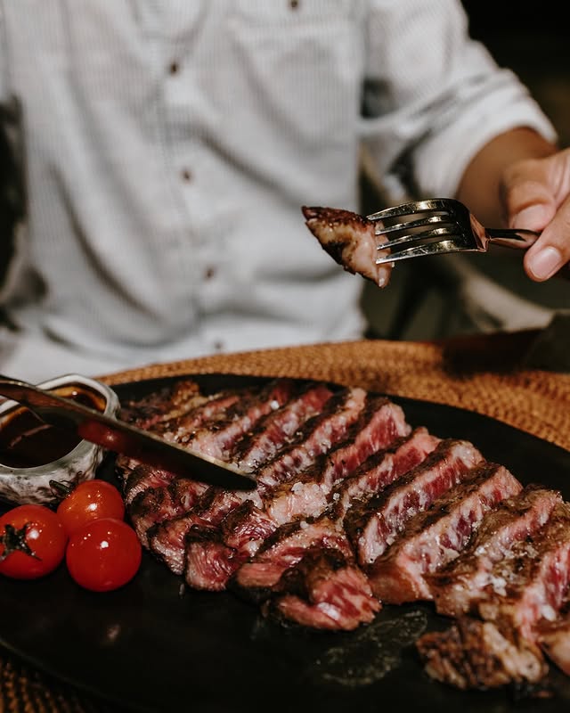 A customer holding a fork with a piece of gourmet steak from a plate full of steak and side of tomatoes