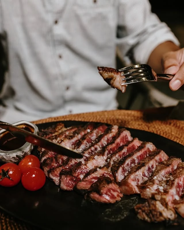 A customer holding a fork with a piece of gourmet steak from a plate full of steak and side of tomatoes